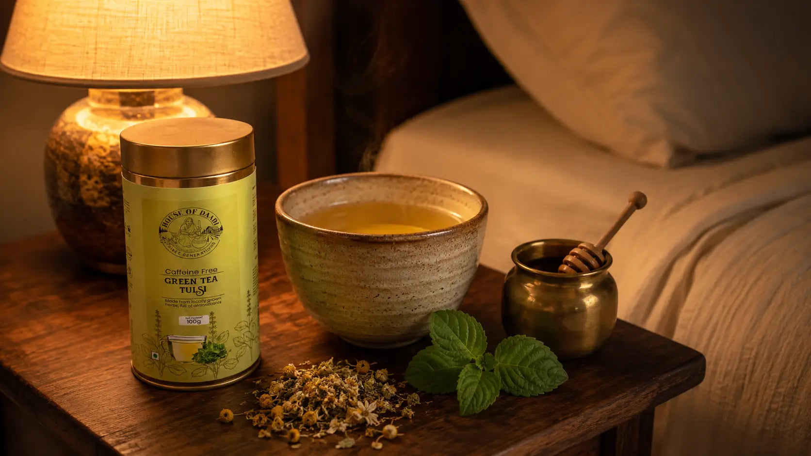 A ceramic cup of steaming chamomile and tulsi herbal tea on a wooden bedside table with dried chamomile flowers, a honey jar, and soft warm lamp light in a dark evening setting
