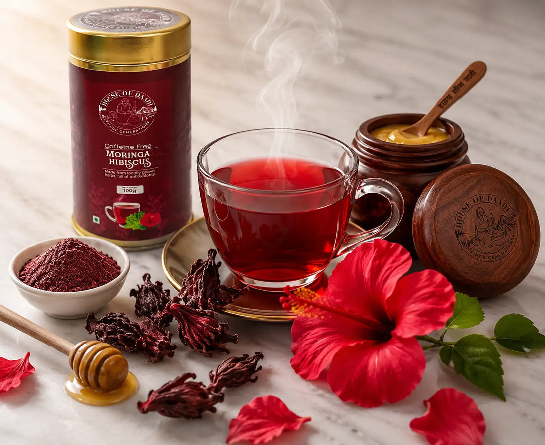 A glass cup of deep ruby-red hibiscus tea with dried hibiscus flowers and fresh hibiscus petals arranged around it on a white marble surface with soft natural light