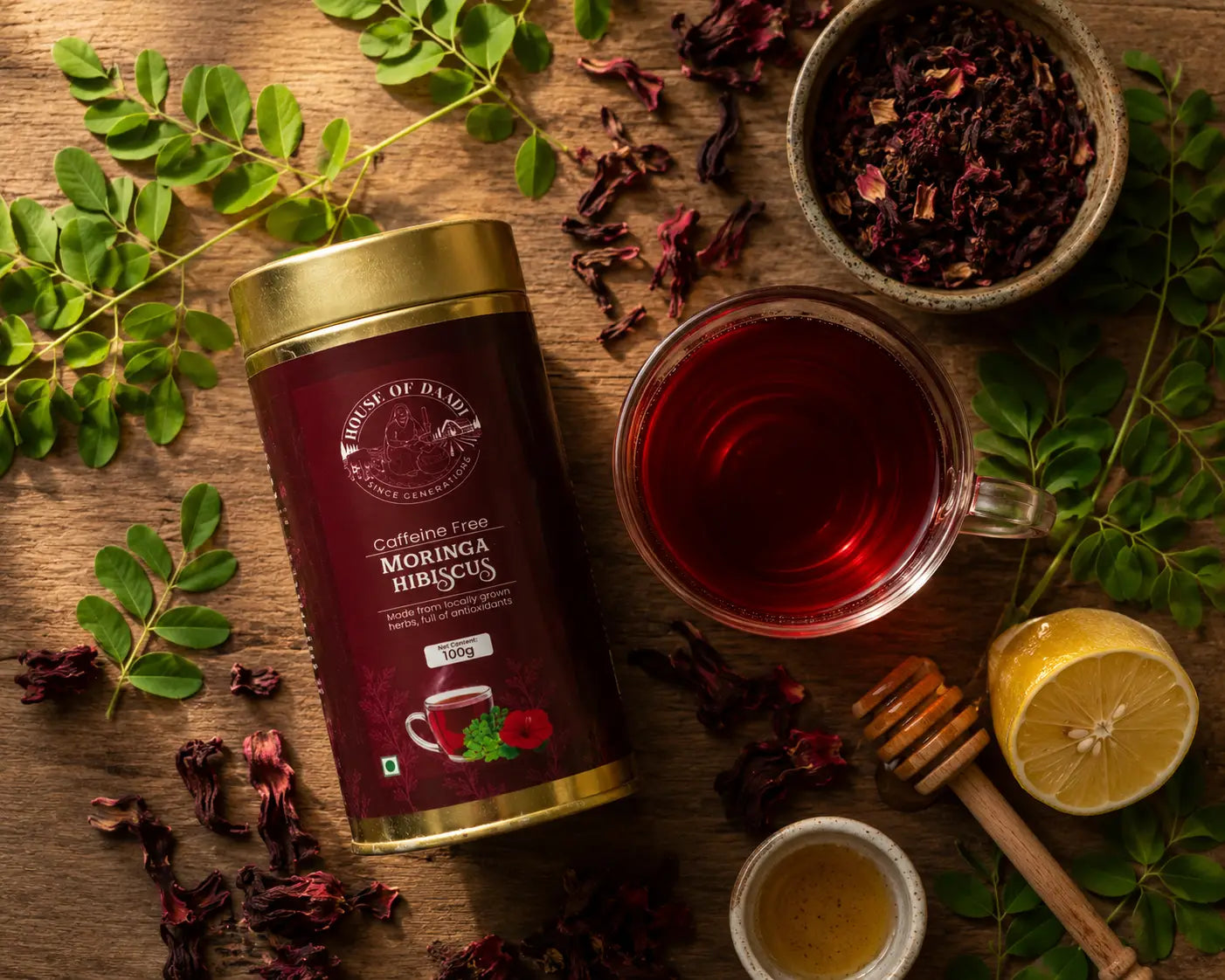 A glass cup of deep ruby-red Moringa Hibiscus Tea surrounded by fresh moringa leaves, dried hibiscus flowers, and a wooden honey dipper on a rustic wooden surface